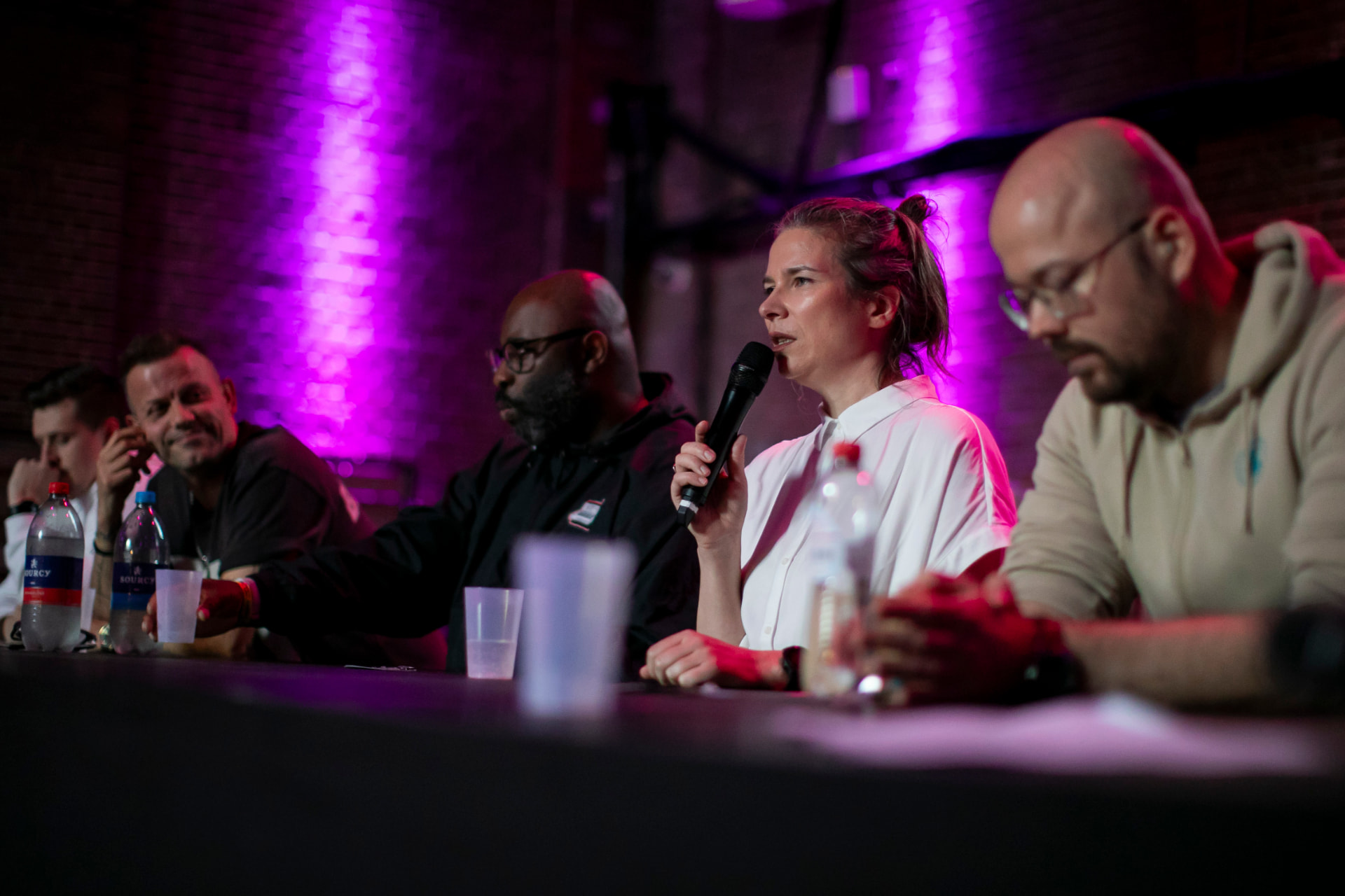 Wide shot of a panel table in purple light, a woman speaking into a microphone