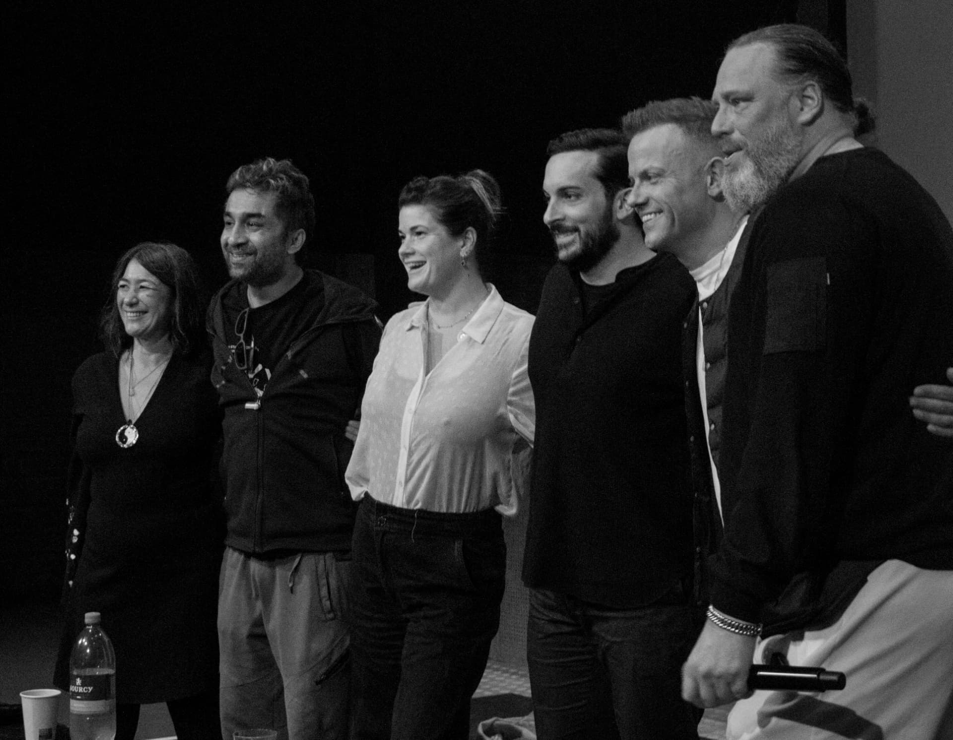 Six contributors smiling together on stage in black and white at the end of the program