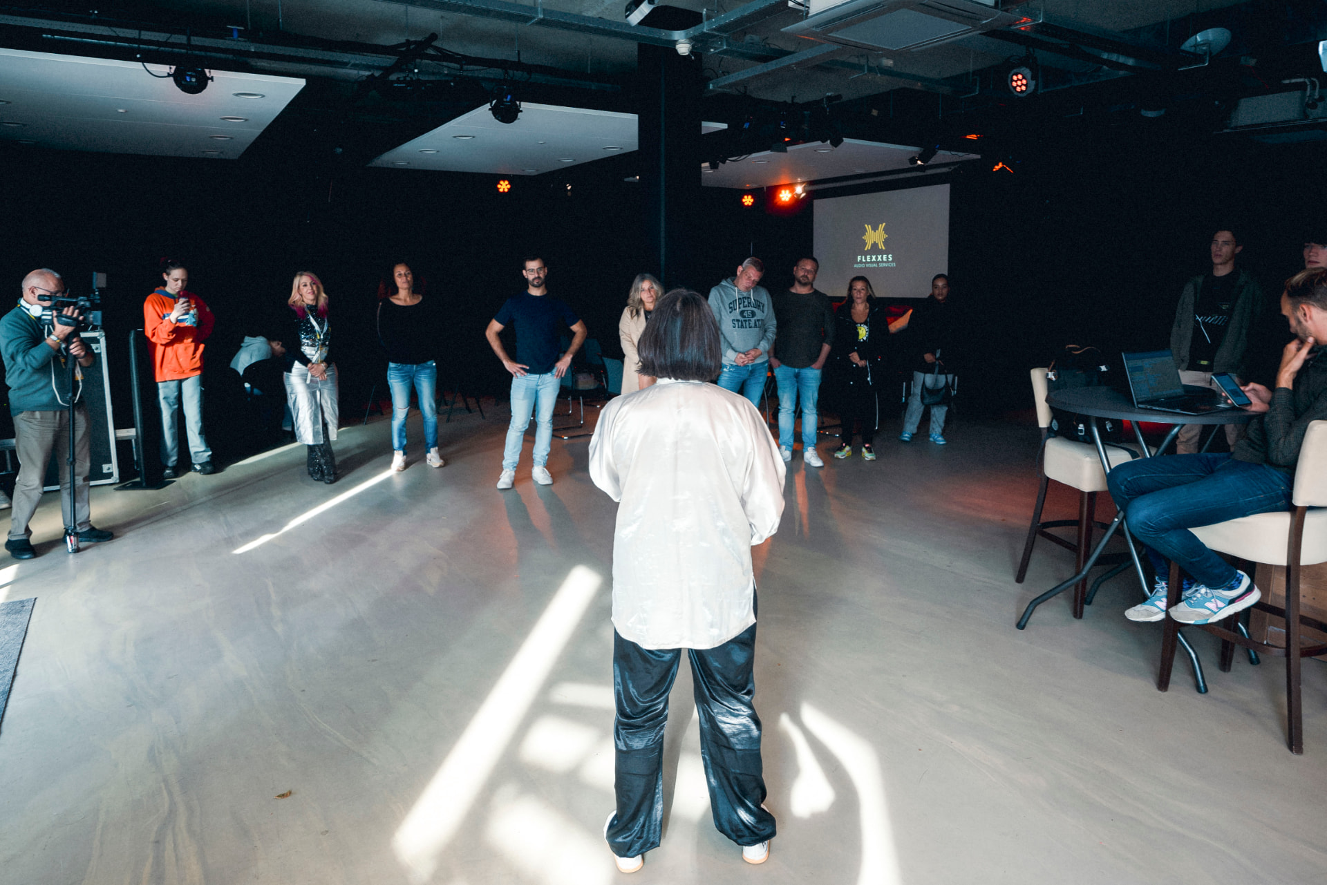 A facilitator leads a standing movement session in a bright room, a small group following from a distance