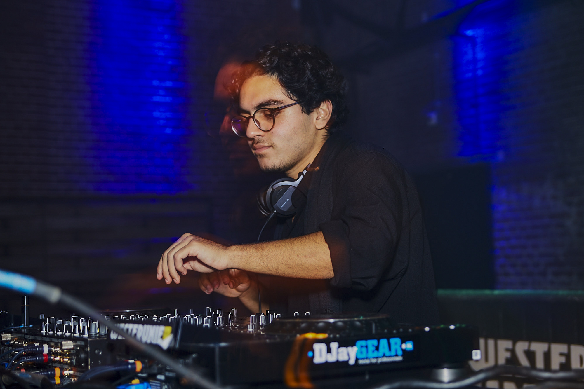 A DJ with glasses and headphones around his neck plays a set behind the decks in blue light