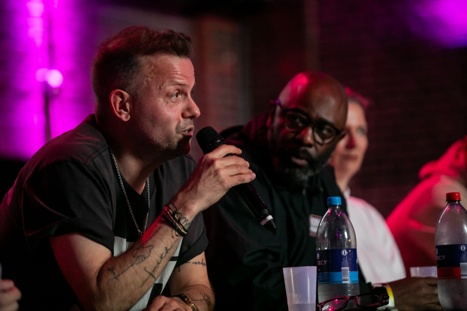 A panel conversation in purple light, a man speaking into a microphone while a listener leans in beside him